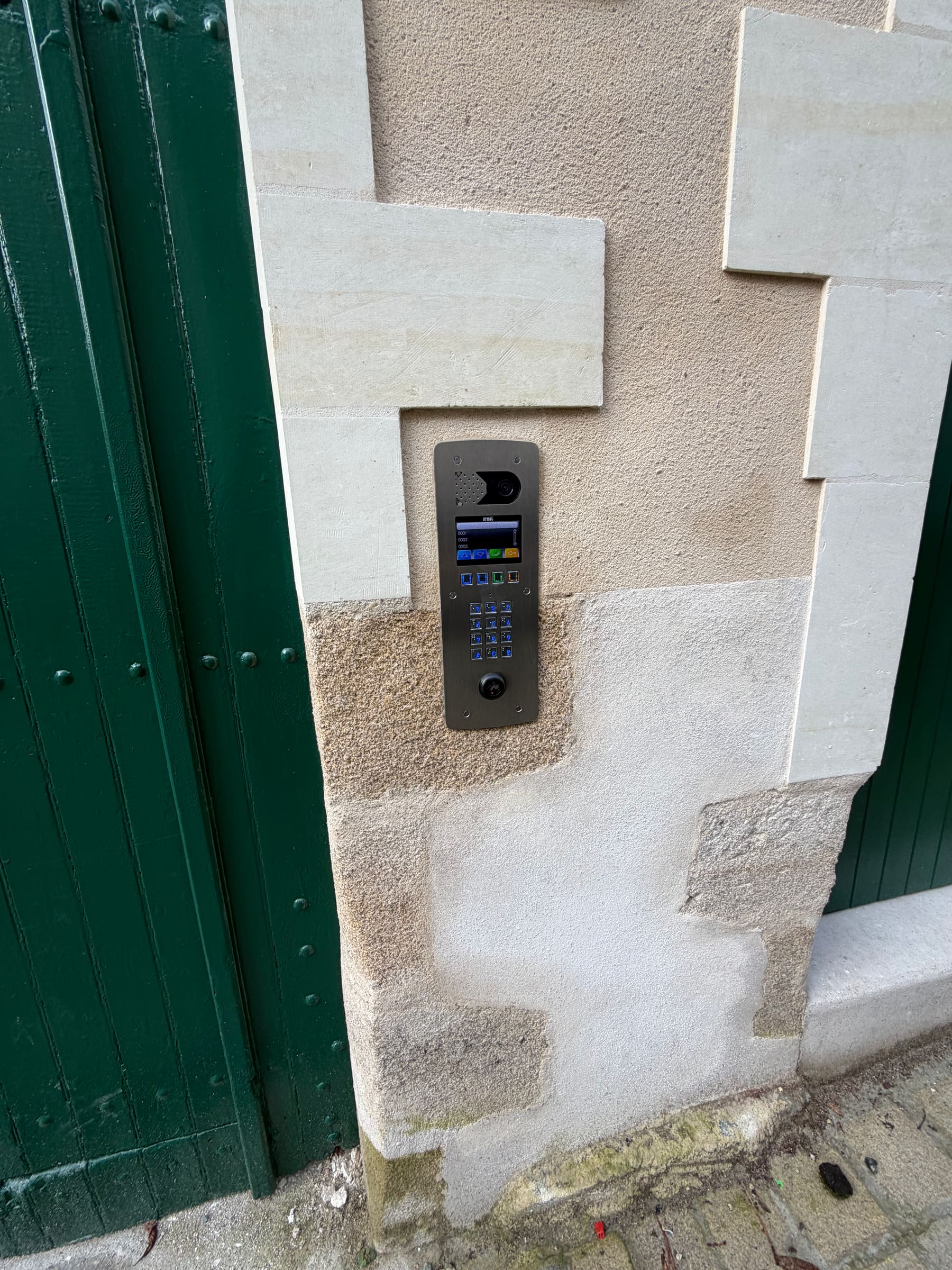 Platine vidéo Urmet à clavier, encastrée dans un pilier en pierre près d’un portail vert