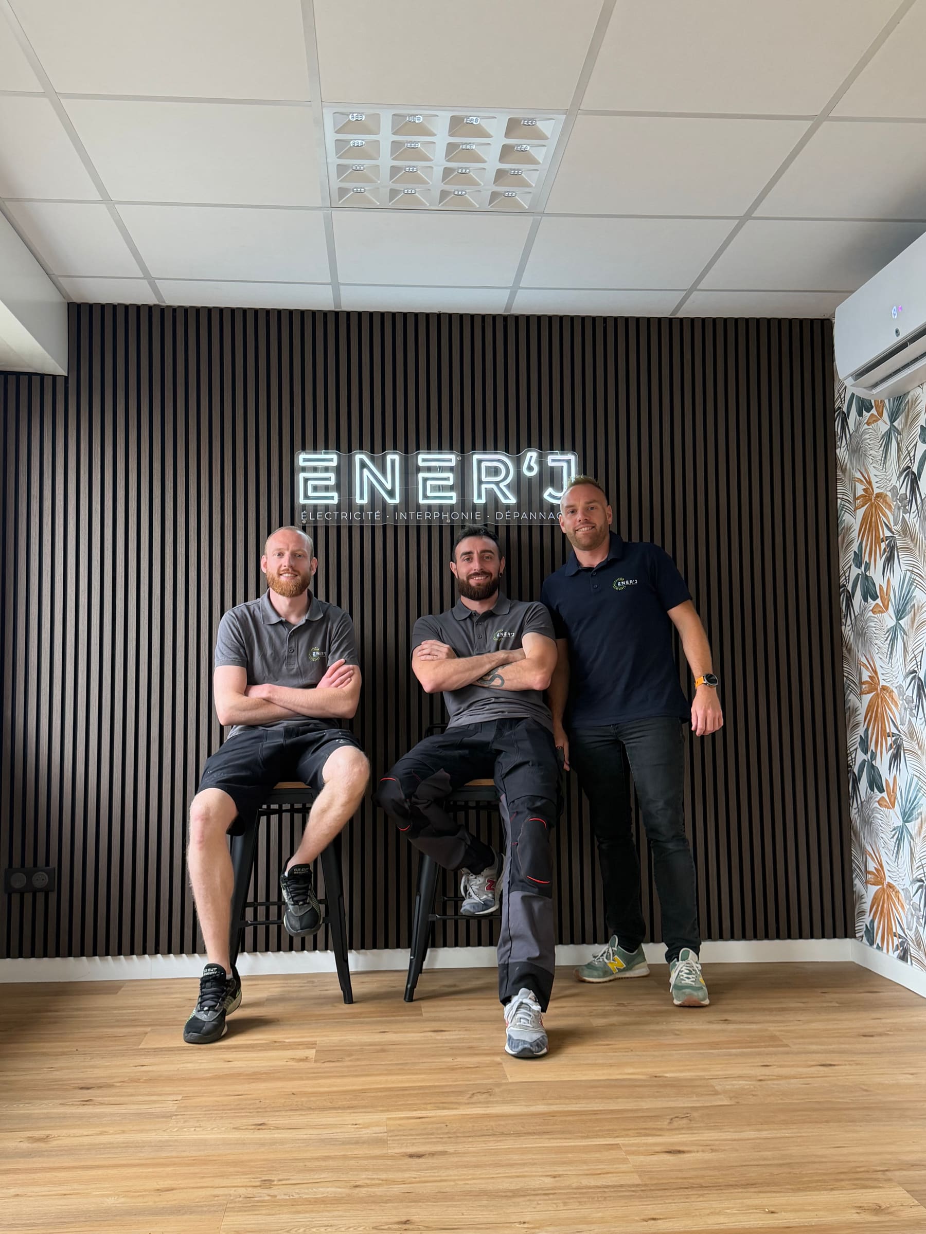 L’équipe ENER'J, trois personnes devant l’enseigne lumineuse, dans leurs locaux